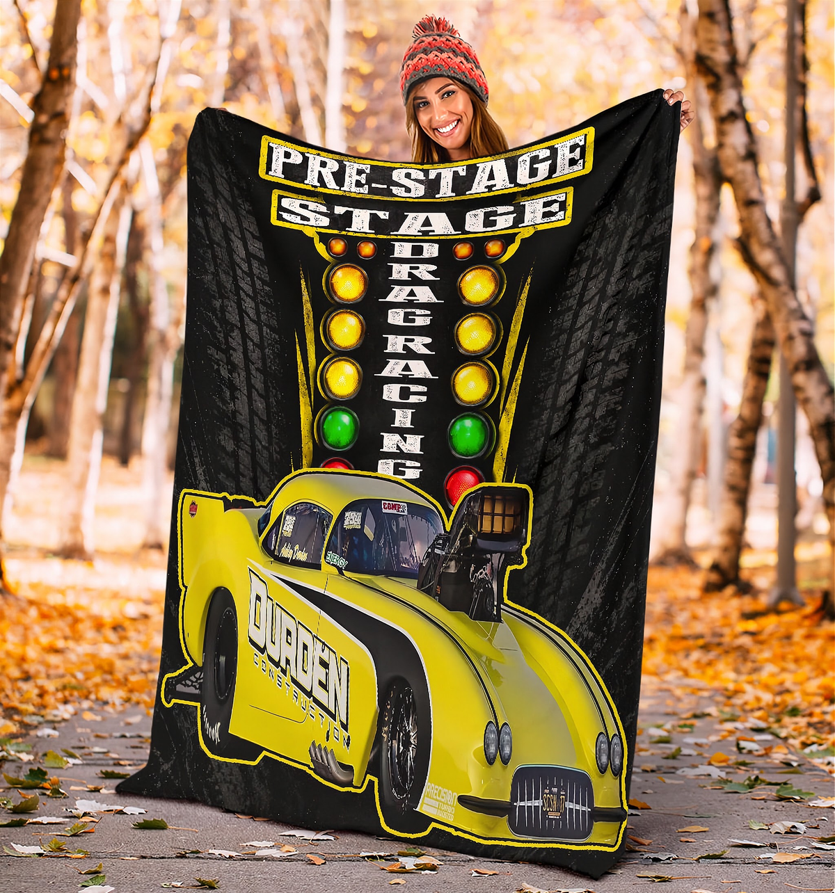 A woman holds up a large black blanket or throw featuring a bright yellow drag racing car and a vertical light bar graphic with the words 'PRE-STAGE DRAG RACING.' The background is an outdoor autumn tree path with fallen leaves.