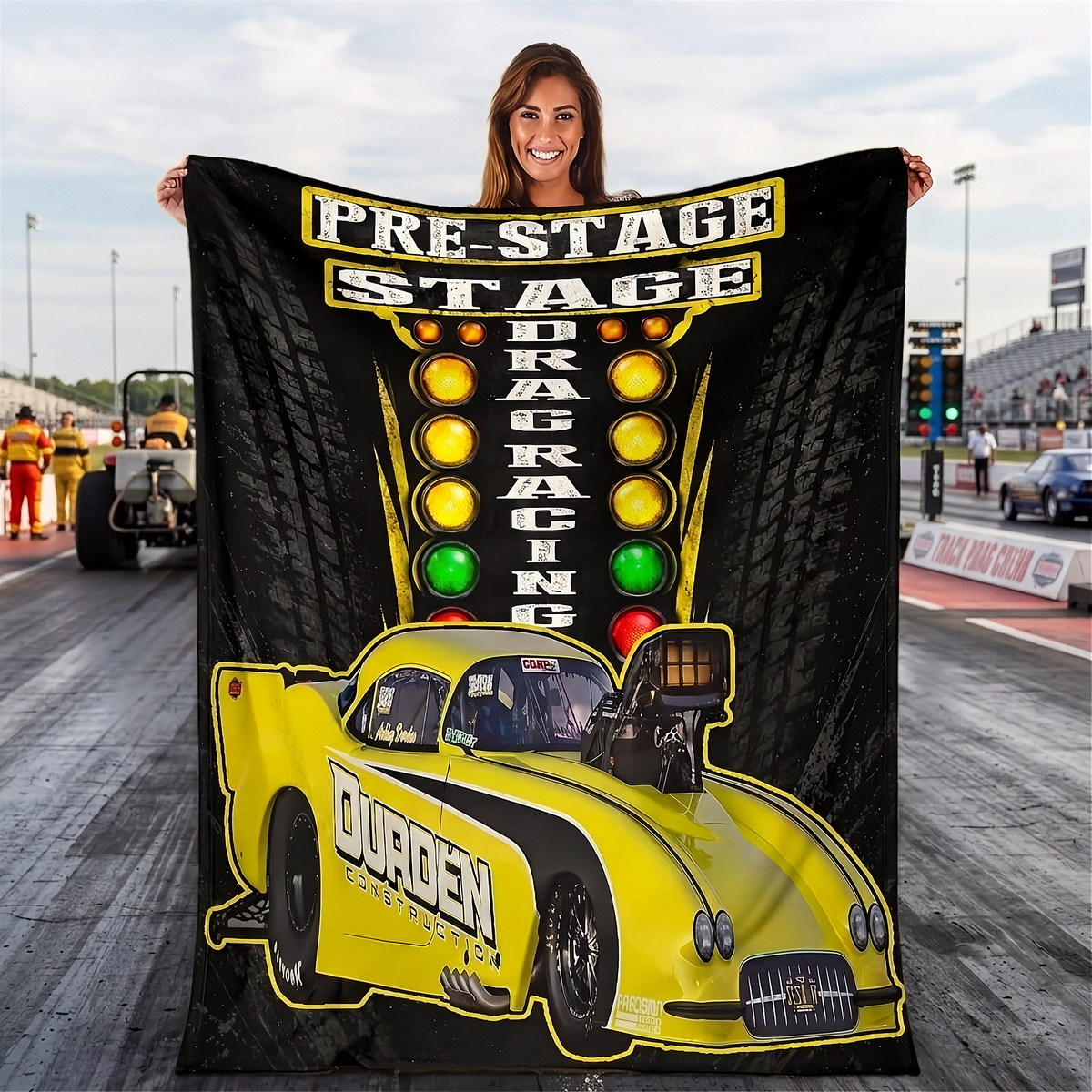 A woman stands on a drag racing track holding up a large black and yellow fleece blanket featuring a drag racer car and the words 'PRE-STAGE DRAG RACING'.