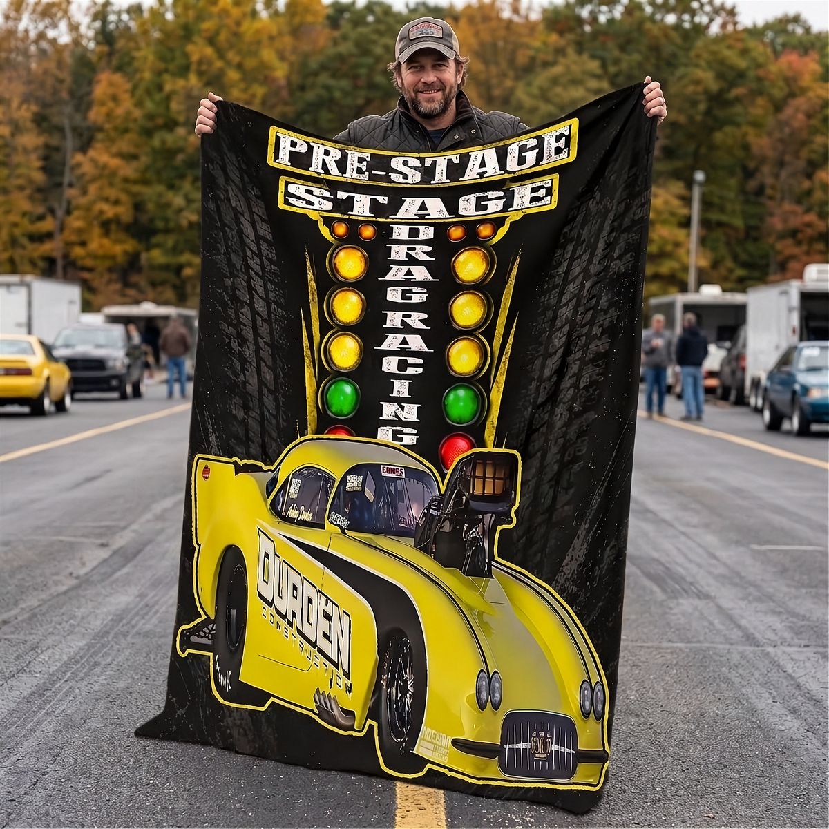 A man stands on an outdoor drag racing track holding up a large black fleece blanket featuring a bright yellow drag racer car and the words 'PRE-STAGE DRAG RACING' with traffic light graphics.