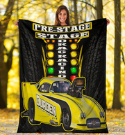 A woman stands outdoors in a wooded area holding up a large black fleece blanket or poster featuring a bright yellow drag racing car and the words 'PRE-STAGE DRAG RACING.' Because a person and a real outdoor environment are visible, this is classified as Lifestyle rather than Product/Packaging, even though the product itself is clearly shown.