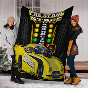Two women holding up a large black fleece blanket featuring a bright yellow drag racing car and a racing light sequence graphic. The scene is set outdoors in front of a house with snow on the ground.