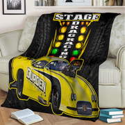 A black throw blanket draped over a light beige sofa in a living room setting, featuring a graphic print of a yellow drag racing car and a vertical marquee reading 'STAGE DRAG RACING'.