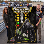 A man and a woman stand on a racetrack holding up a large black fleece blanket or throw. The blanket features a graphic of a motorcycle racer in a black suit with green and white accents, a vertical row of colored lights, and the words 'PRE-STAGE DRAG RACING' and 'POWER EDGE'.