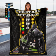 A woman stands on a drag racing track holding up a large black fleece blanket or throw featuring a racing motorcycle, driver, tire tread graphics, and the words 'PRE-STAGE DRAG RACING' with colored light buttons.
