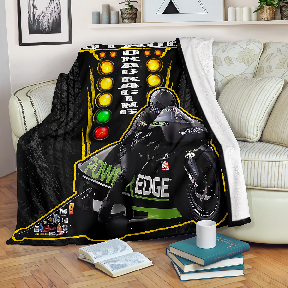 A black throw blanket featuring a racing graphic with a motorcycle, driver, and 'Drag Racing' text, draped over a cream-colored sofa in a living room setting.