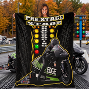 A woman stands on a racetrack holding a large black fleece blanket printed with a racing graphic. The blanket features the text 'PRE-STAGE DRAG RACING' in yellow and white, a motorcycle racer illustration, and green and yellow accents.