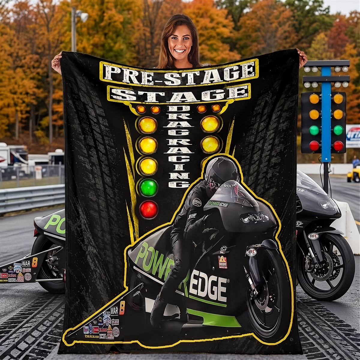 A woman stands on a racetrack holding a large black fleece blanket printed with a racing graphic. The blanket features the text 'PRE-STAGE DRAG RACING' in yellow and white, a motorcycle racer illustration, and green and yellow accents.