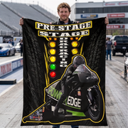 A man stands on a racetrack holding up a large black custom blanket or poster featuring a drag racing theme with a motorcycle racer, tire textures, and colored light bulbs.