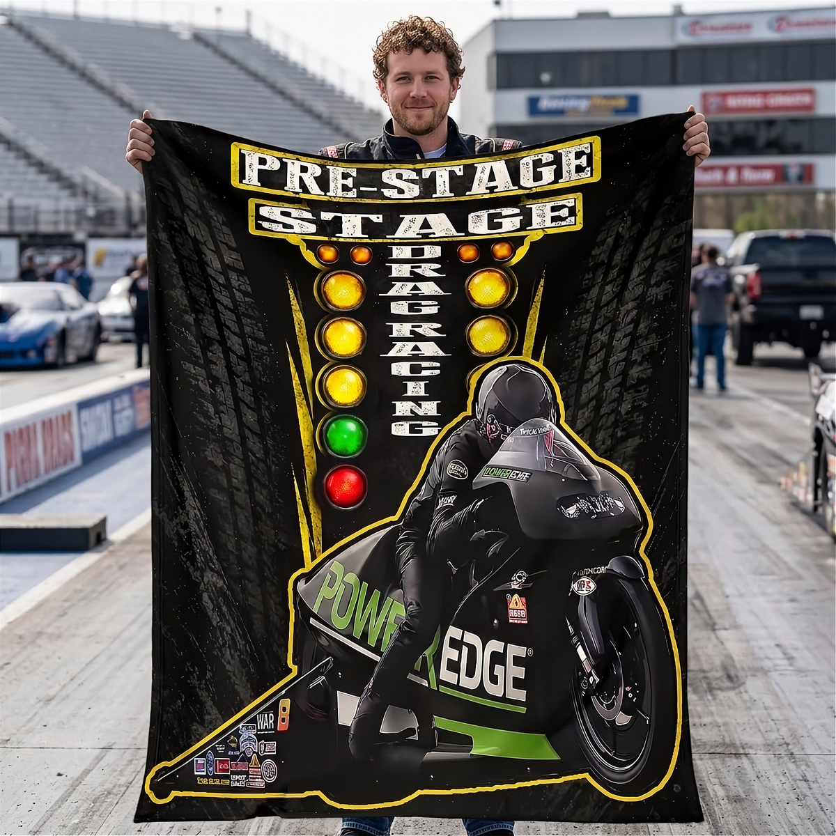 A man stands on a racetrack holding up a large black custom blanket or poster featuring a drag racing theme with a motorcycle racer, tire textures, and colored light bulbs.