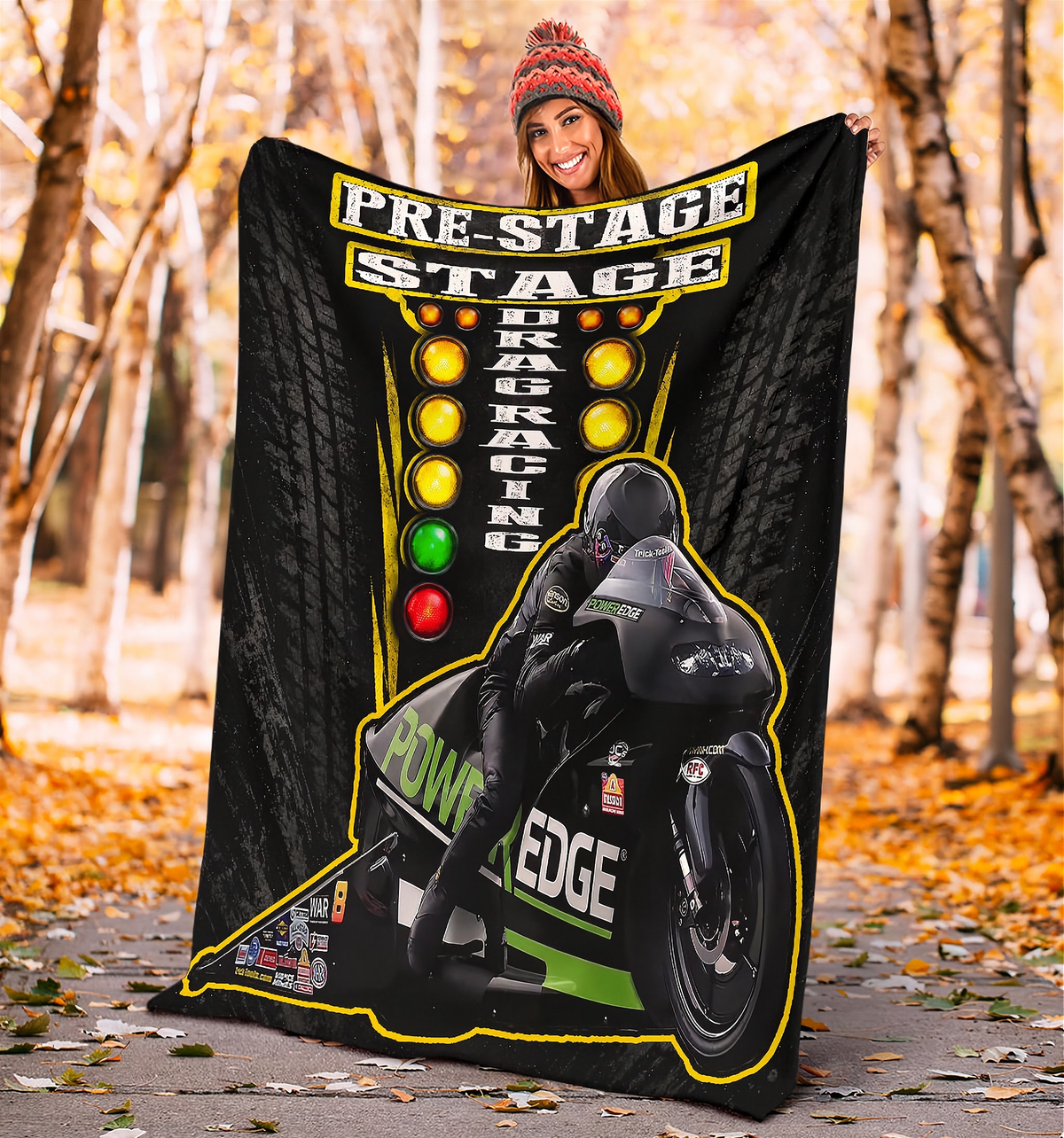 A person standing outdoors in a wooded park holding up a large black fleece blanket or throw. The blanket features a racing graphic with the words 'PRE-STAGE DRAG RACING,' a motorcycle illustration, and illuminated traffic-light style circles in yellow, amber, green, and red.
