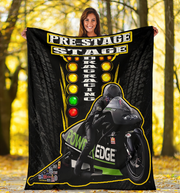 A woman stands outdoors holding up a large black fleece blanket or poster featuring drag racing graphics and a motorcycle racer. The design includes the words 'PRE-STAGE STAGE DRAG RACING' and 'POWER EDGE' with yellow, green, and red racing light elements and tire tread patterns in the background.