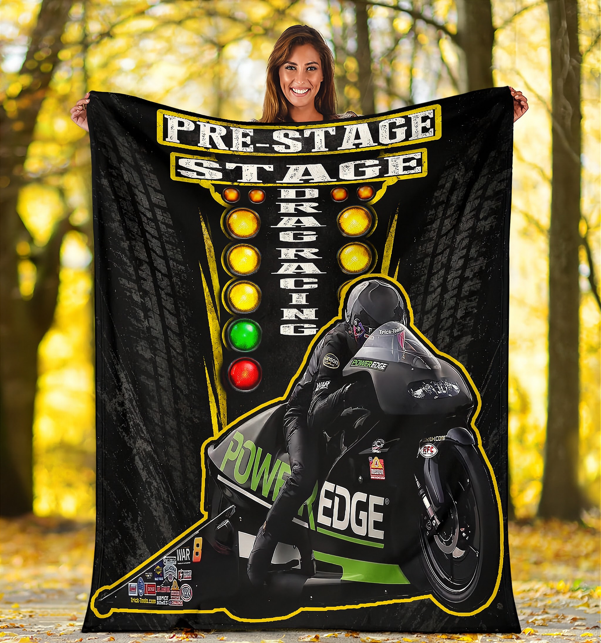 A woman stands outdoors holding up a large black fleece blanket or poster featuring drag racing graphics and a motorcycle racer. The design includes the words 'PRE-STAGE STAGE DRAG RACING' and 'POWER EDGE' with yellow, green, and red racing light elements and tire tread patterns in the background.