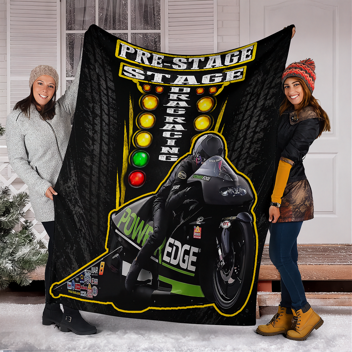 Two women standing outdoors in front of a house door holding up a large black fleece blanket or throw featuring a drag racing graphic with a motorcycle racer, the words 'PRE-STAGE DRAG RACING,' and colored light buttons.