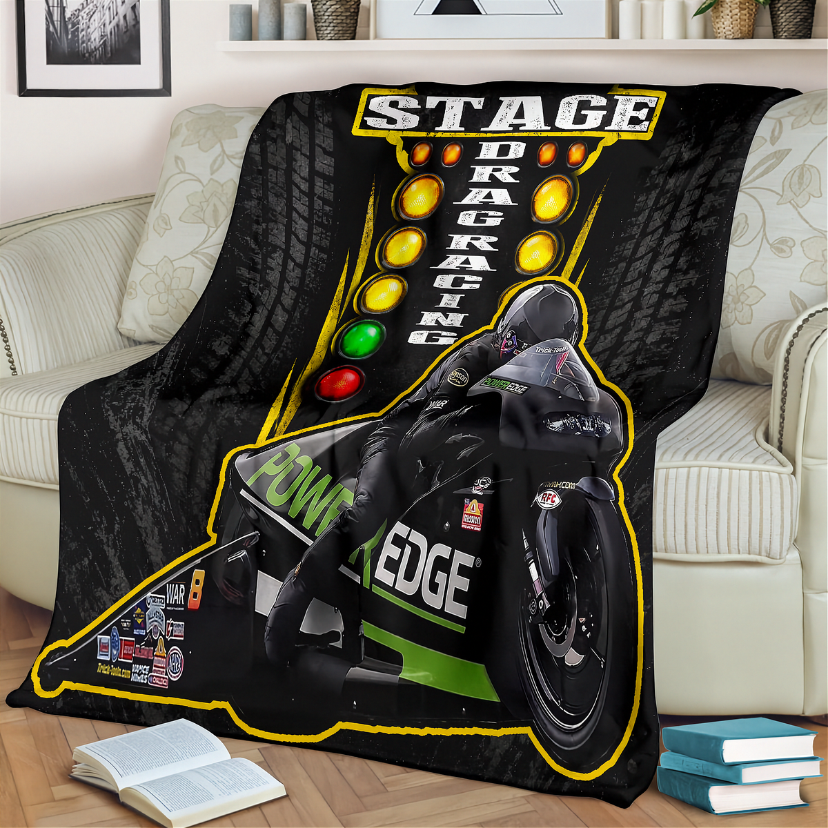 A black throw blanket draped over a light beige sofa in a living room setting, featuring a graphic print of a drag racing motorcycle with 'STAGE DRAG RACING' text and yellow light bulbs.
