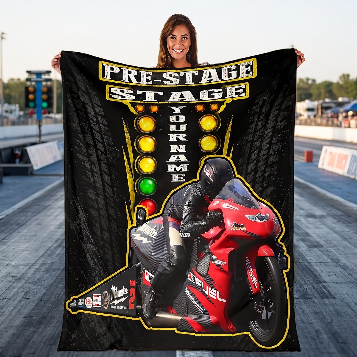 A woman stands on a racetrack holding up a large custom-printed black blanket or throw featuring a red motorcycle, racing graphics, and the text 'PRE-STAGE STAGE YOUR NAME'.