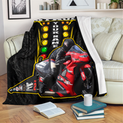 A custom motorcycle-themed throw blanket draped over a sofa in a living room setting. The blanket features a black and red racing motorcycle graphic with a racer, yellow circular lights, and customizable text reading 'YOUR NAME' and 'SPARE THE LIGHT'.