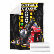 A custom motorcycle-themed fleece blanket shown flat with a folded white sherpa backing visible at the top left corner, and two rolled white fleece ends at the bottom right. The blanket features a black background with tire tread graphics, a red sportbike, a motorcycle racer in black leather gear, and yellow light bulbs with the text 'MOTORCYCLE RACE' and 'YOUR NAME'.