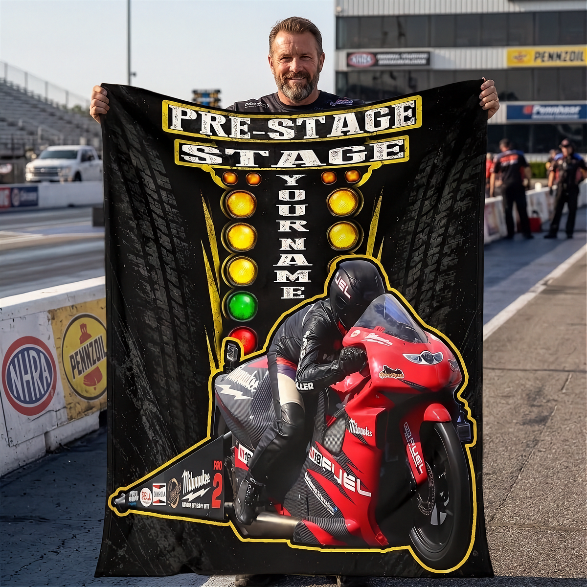 A man stands on a racetrack holding up a large black custom fleece blanket or poster featuring a red racing motorcycle, a rider, and a checkered light sequence with the text 'PRE-STAGE STAGE YOUR NAME'.