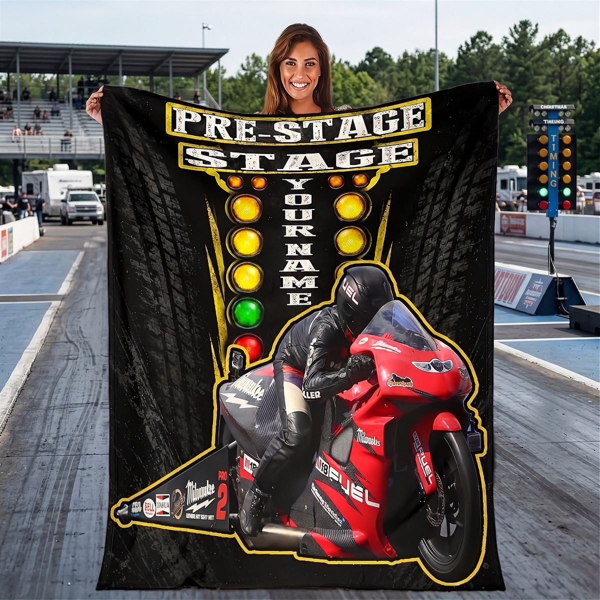 A woman stands on a racetrack holding a large custom-printed blanket or poster. The print features a black background with tire tracks, yellow racing lights, the text 'PRE-STAGE STAGE YOUR NAME', and a graphic of a racer on a red motorcycle.