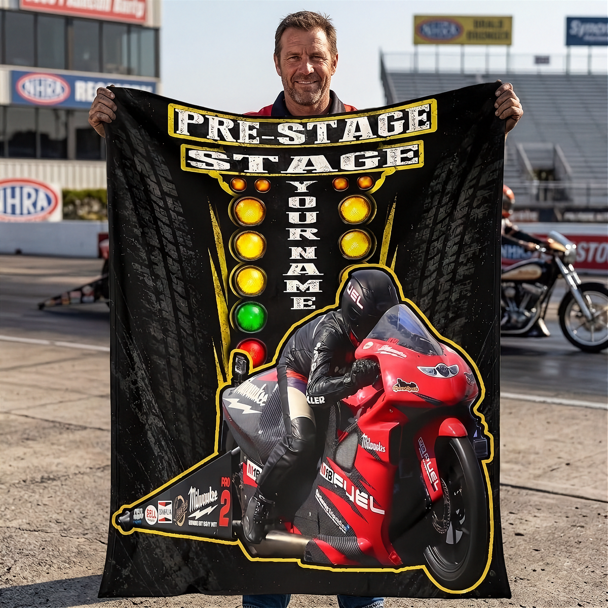 A man standing on a racetrack holding up a large custom-printed black blanket or poster featuring a red sport motorcycle, a racer in black gear, and the text 'PRE-STAGE STAGE' with a customizable name column.