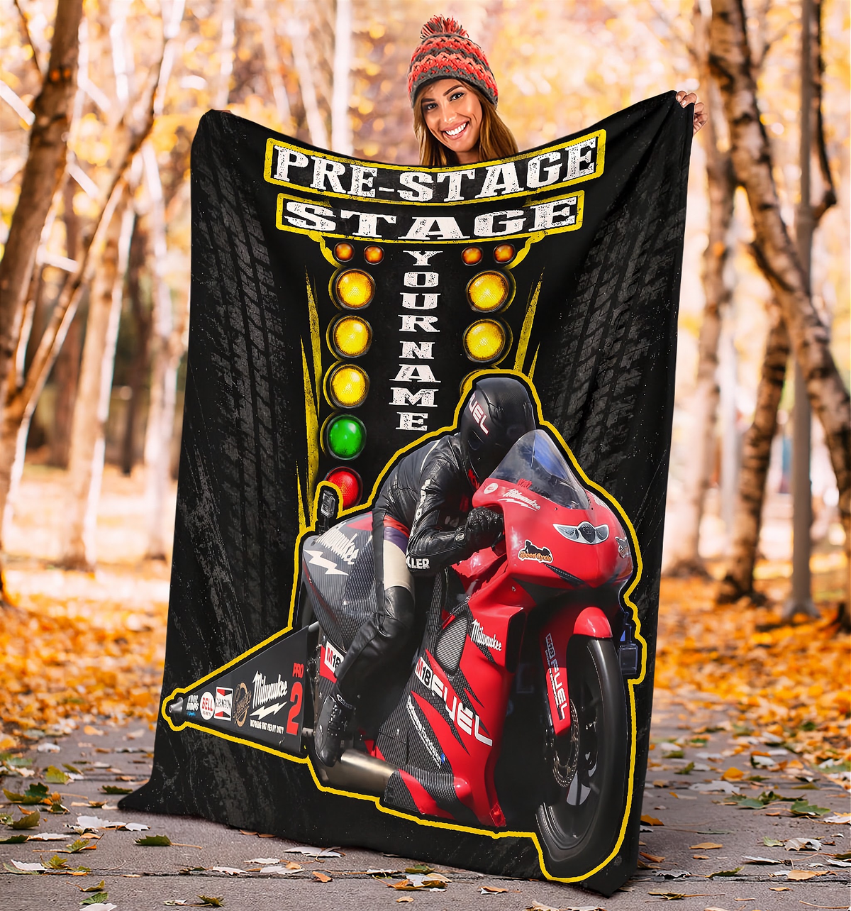 A woman wearing a red knit beanie holds up a large custom motorcycle-themed blanket or poster in an outdoor autumn setting with fallen leaves and trees in the background. The product features a black tire-pattern background, yellow 'PRE-STAGE STAGE' and 'YOURNAME' text, and a graphic of a rider on a red and black motorcycle.