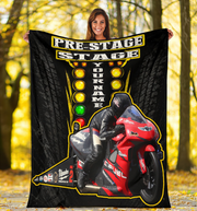 A woman stands outdoors in a wooded area holding up a large custom-printed blanket or poster. The product features a black background with tire tracks, a red and black racing motorcycle, a rider in black gear, and yellow text reading 'PRE-STAGE STAGE' and 'YOUR NAME'.