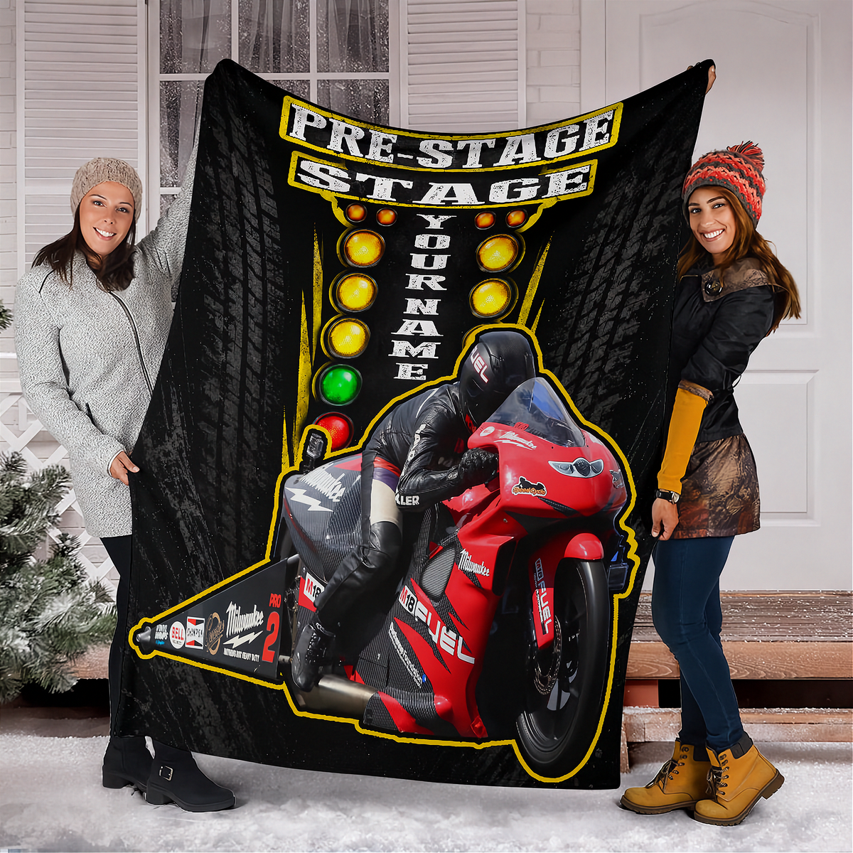 A large black custom motorcycle-themed blanket or tapestry is held up by two smiling women standing in front of a house. The blanket features a red racing motorcycle, yellow track lights, and the text 'PRE-STAGE STAGE' and 'YOUR NAME'.