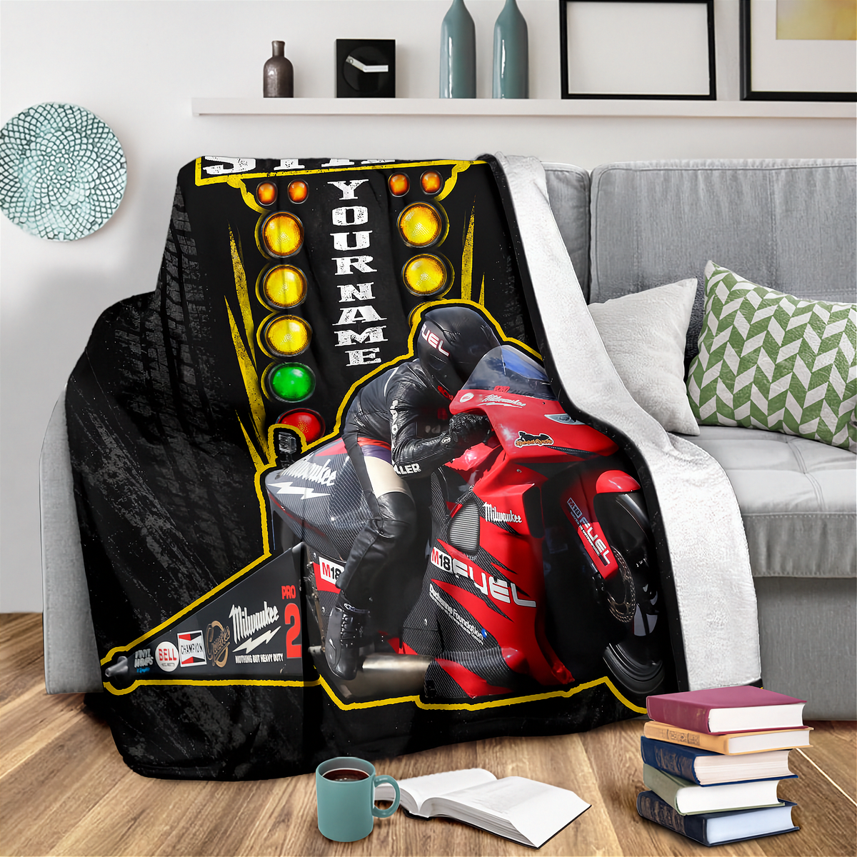A fleece throw blanket featuring a custom racing design with a red motorcycle, a racer in black gear, and a traffic light pattern, draped over a gray sofa in a living room setting.