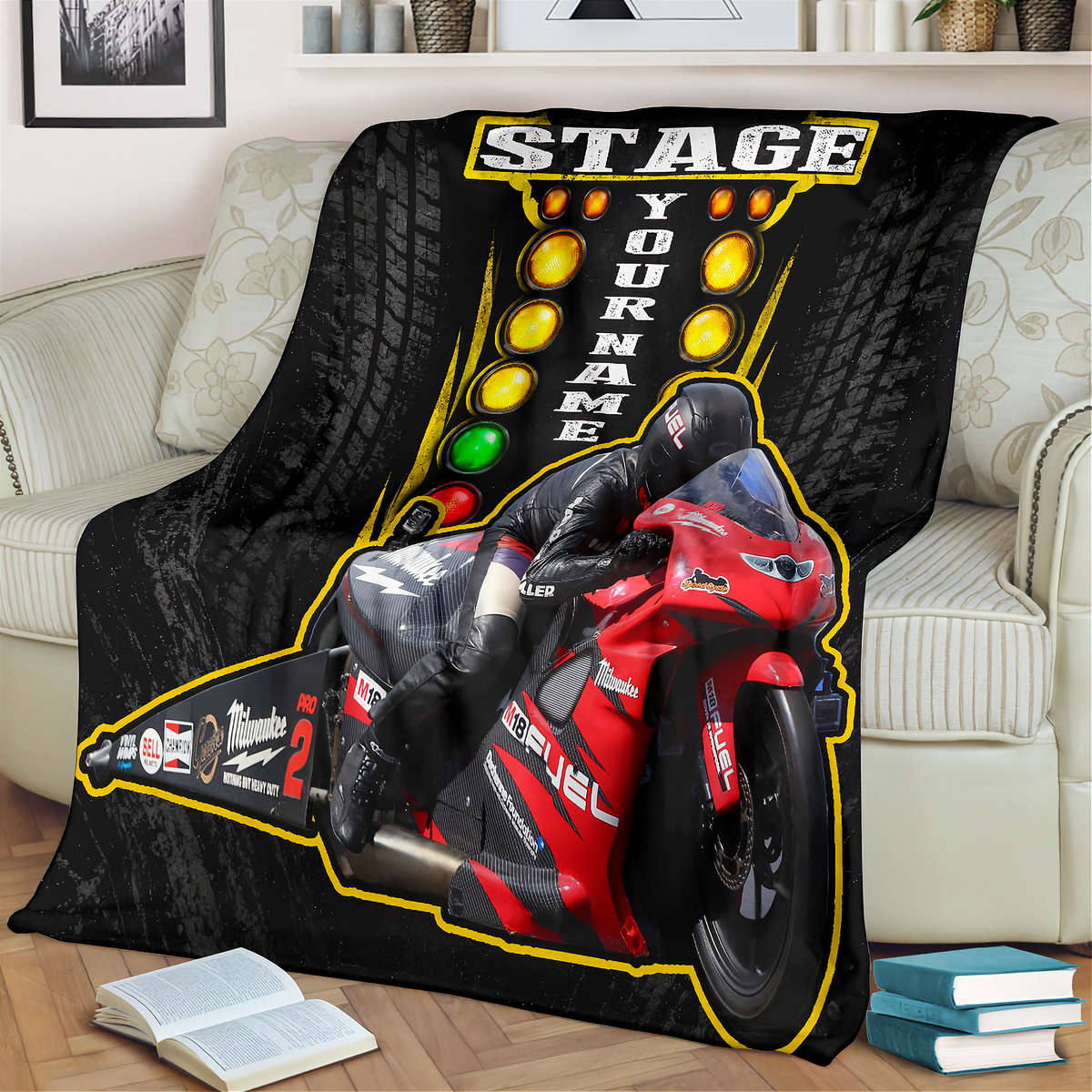 A custom printed fleece throw blanket draped over a beige sofa in a living room setting. The design features a red and black racing motorcycle with a rider, a yellow and black track light sequence, and the customizable text 'STAGE YOUR NAME'.