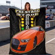 A woman standing on a racetrack holds up a large black printed blanket or tapestry featuring an orange race car and the words 'PRE-STAGE DRAG RACING' with yellow and green lights. Other racetrack elements and people are visible in the background.