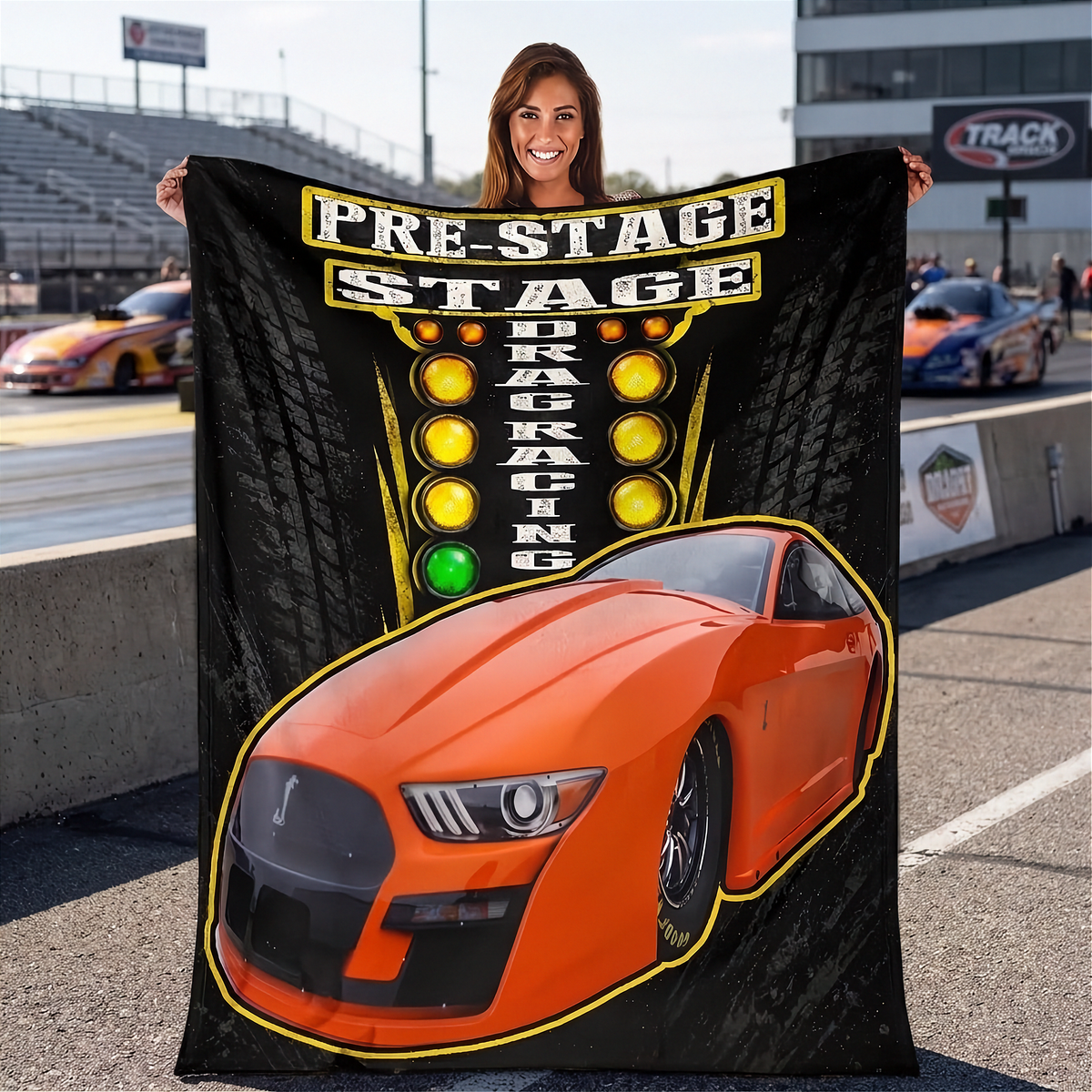 A woman standing on a racetrack holds up a large black printed blanket or tapestry featuring an orange race car and the words 'PRE-STAGE DRAG RACING' with yellow and green lights. Other racetrack elements and people are visible in the background.
