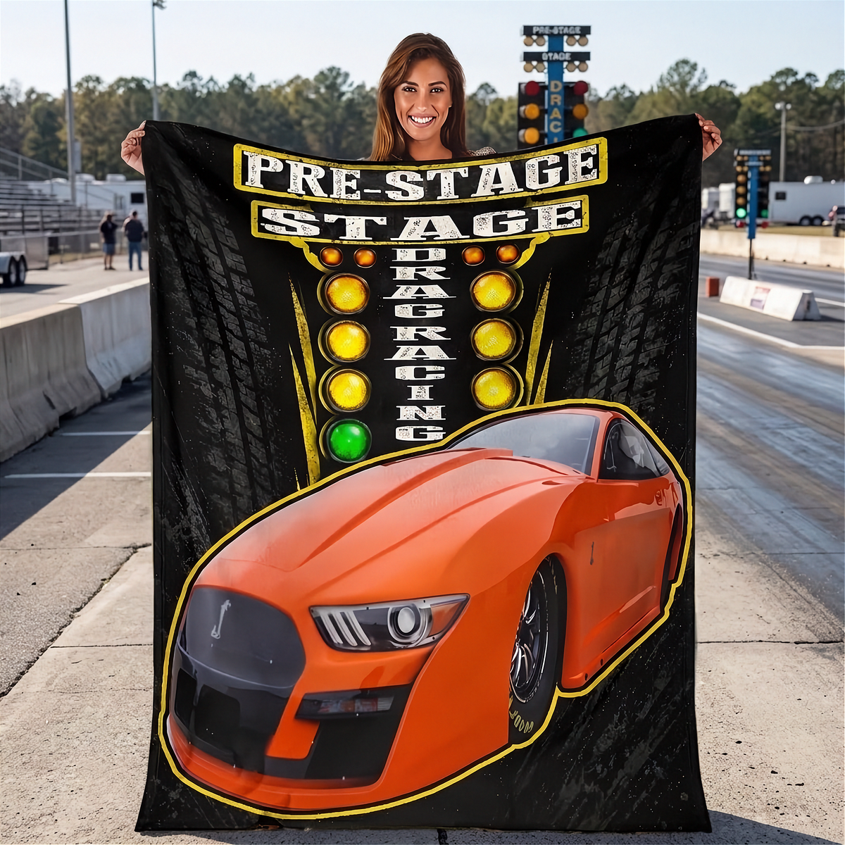 A woman standing on a drag racetrack holds up a large black printed blanket or tapestry featuring an orange race car and the words 'PRE-STAGE DRAG RACING' with yellow and green lights. Other racetrack elements and people are visible in the background.