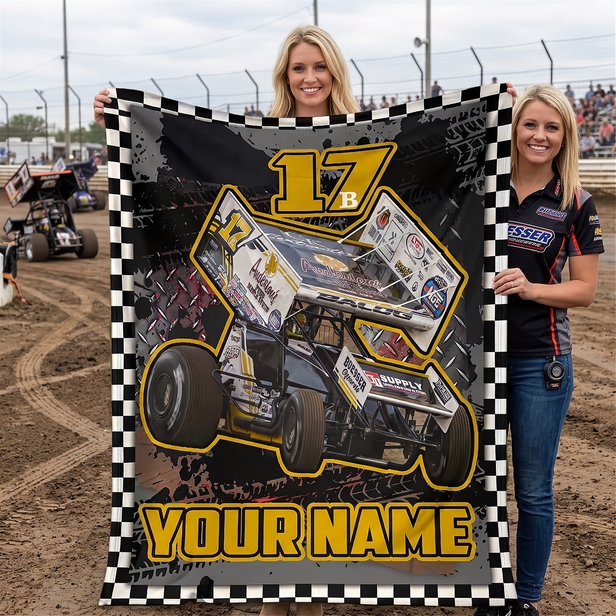 Two women standing on a dirt racetrack holding up a large customizable blanket featuring a race car graphic, the number 17, and 'YOUR NAME' text.
