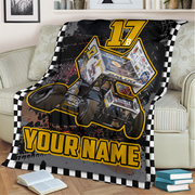 A custom-printed racing blanket draped over a beige sofa in a living room setting. The blanket features a black and white checkered border, a graphic of a sprint car with sponsor decals, the number '17', and the text 'YOUR NAME' in bold yellow letters.