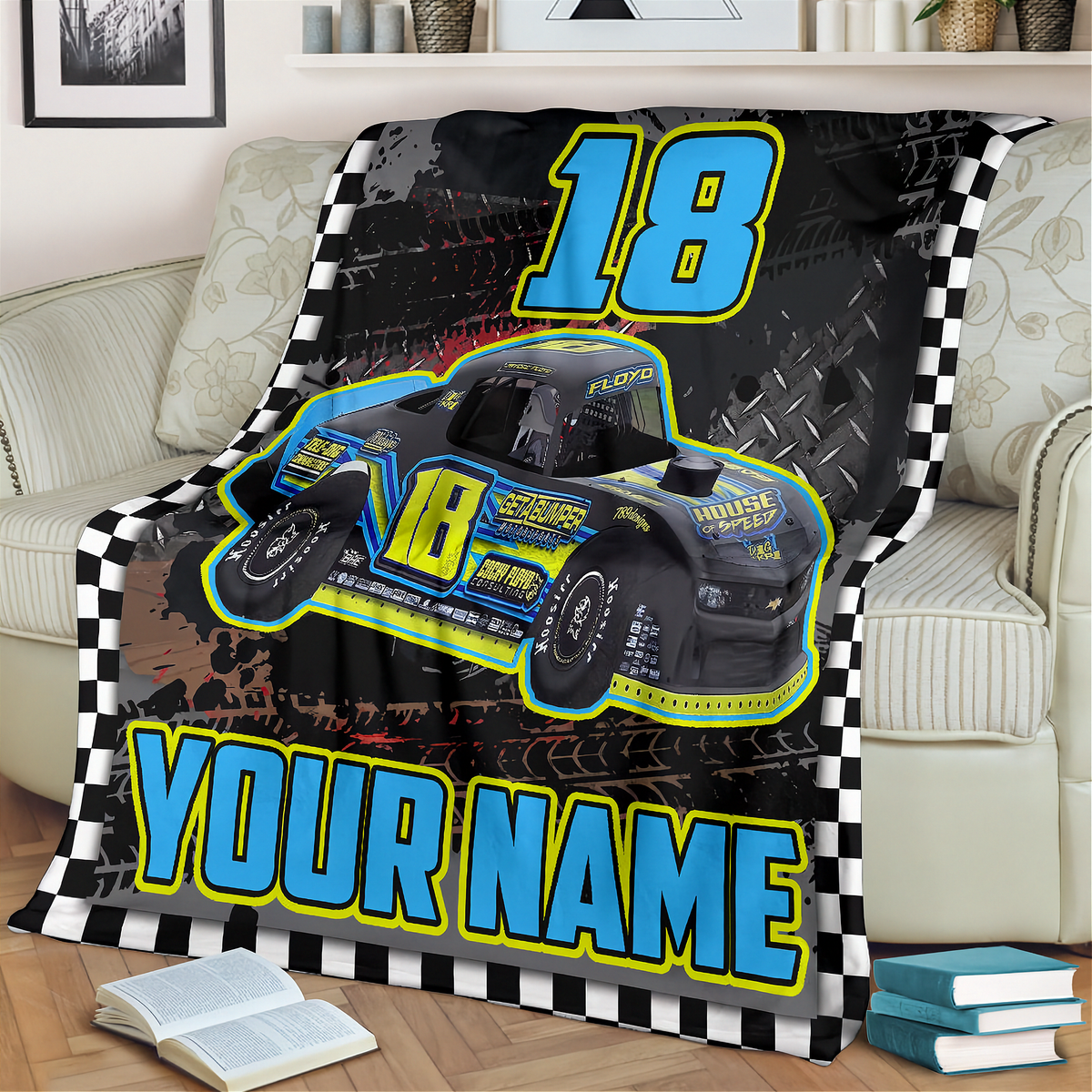 A custom racing-themed blanket draped over a beige sofa in a living room setting. The blanket features a black background with a checkered border, a blue and yellow street stock race car graphic, the number '18', and the text 'YOUR NAME'.