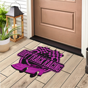 A rectangular doormat with a purple and black drift track racing design, placed on a light wood porch floor in front of a brown door, with a lantern and plant nearby.