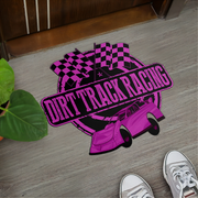 A rectangular floor mat with a 'Dirt Track Racing' graphic in vibrant pink and black, placed on a gray wood floor next to a green plant and near a person's shoes.