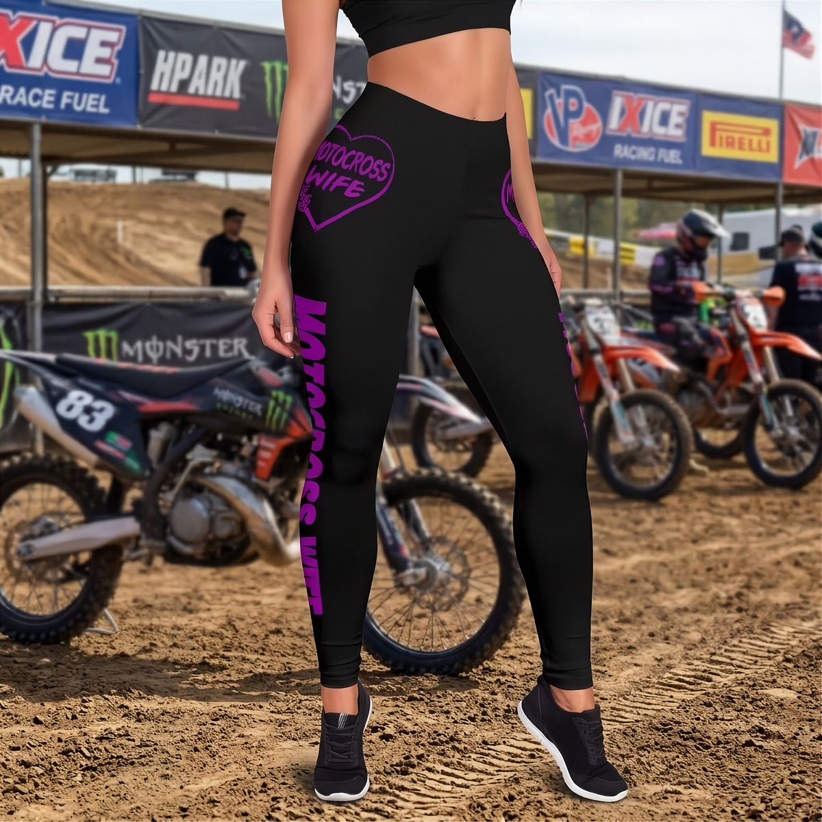 A person wearing black high-waisted leggings with bright purple 'Motorcross Wife' text and a heart graphic, standing in a dirt motocross track setting with bikes and trackside banners in the background.