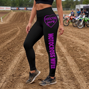 Black high-waisted motocross leggings with purple 'MOTOCROSS WIFE' text graphics, shown on a model standing on a dirt track with motocross bikes in the background.
