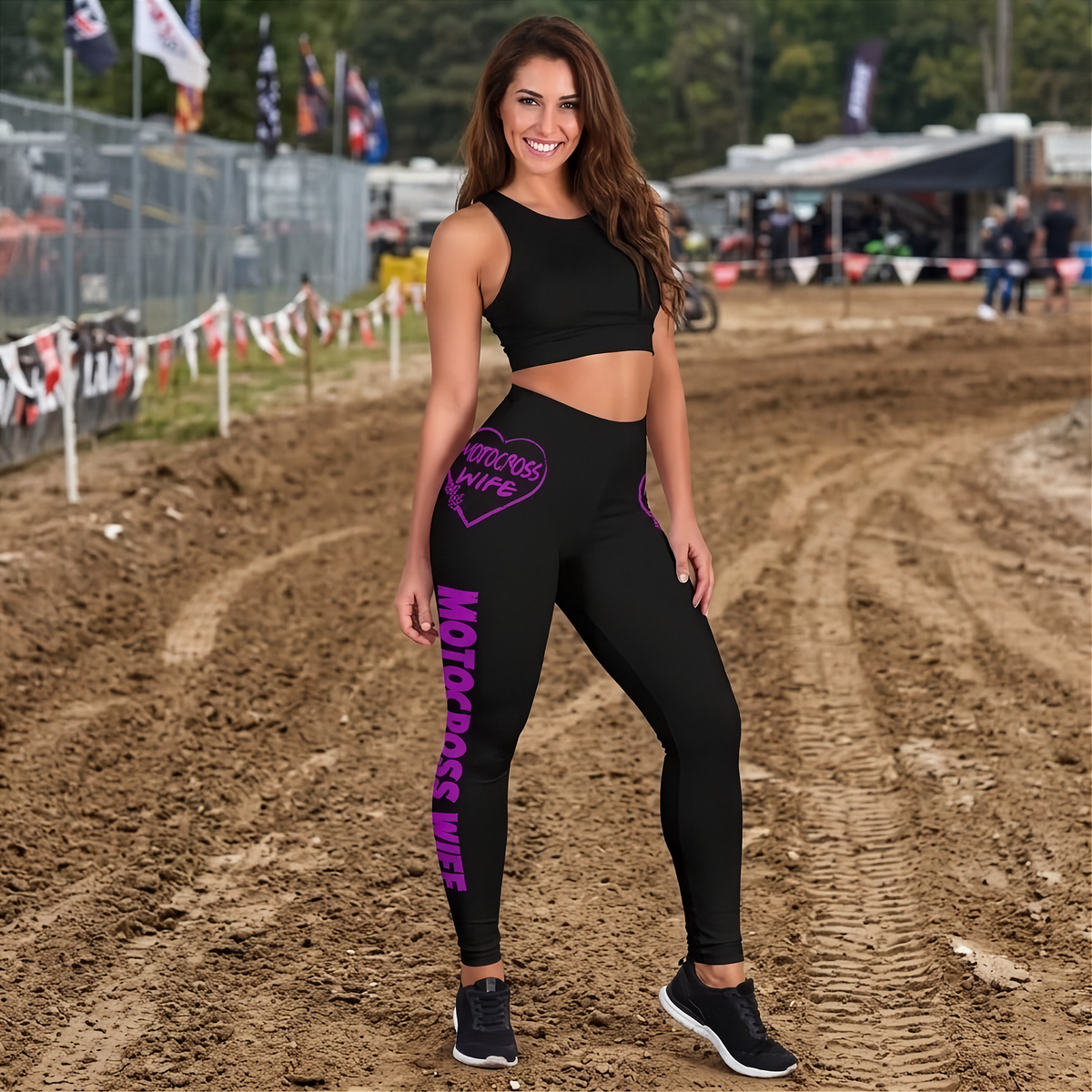 A woman stands on a dirt motocross track wearing a matching black athletic set with bright purple 'MOTORCROSS WIFE' text graphics on the leggings and side of the crop top.