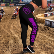 Black athletic leggings with purple 'MOTOCROSS WIFE' text and a heart logo on the back, shown on a motocross track with riders and hay bales in the background.