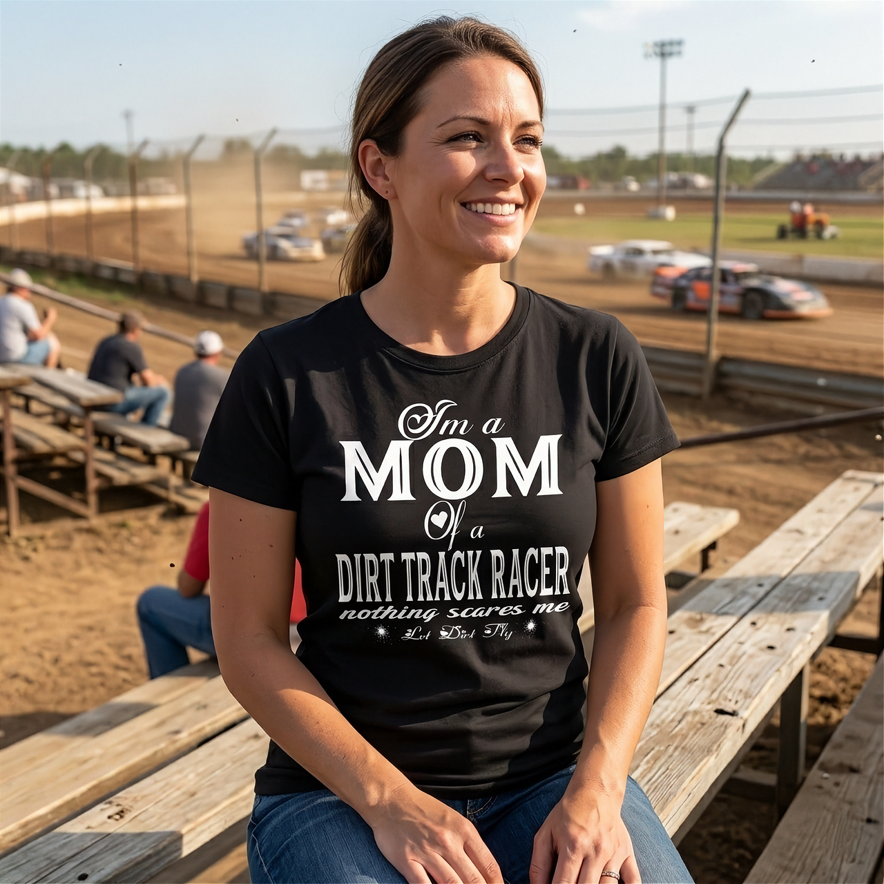 Premium black apparel mockup featuring a woman sitting on the wooden grandstand bleachers at a dirt track arena, wearing a soft custom t-shirt with a bold white graphic that reads "I'm a MOM of a DIRT TRACK RACER nothing scares me".