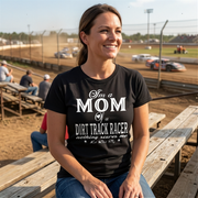 Premium black apparel mockup featuring a woman sitting on the wooden grandstand bleachers at a dirt track arena, wearing a soft custom t-shirt with a bold white graphic that reads "I'm a MOM of a DIRT TRACK RACER nothing scares me".