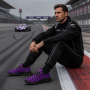 Man sitting on a race track wearing black outfit and purple checkered shoes with a blurred racing car in the background.