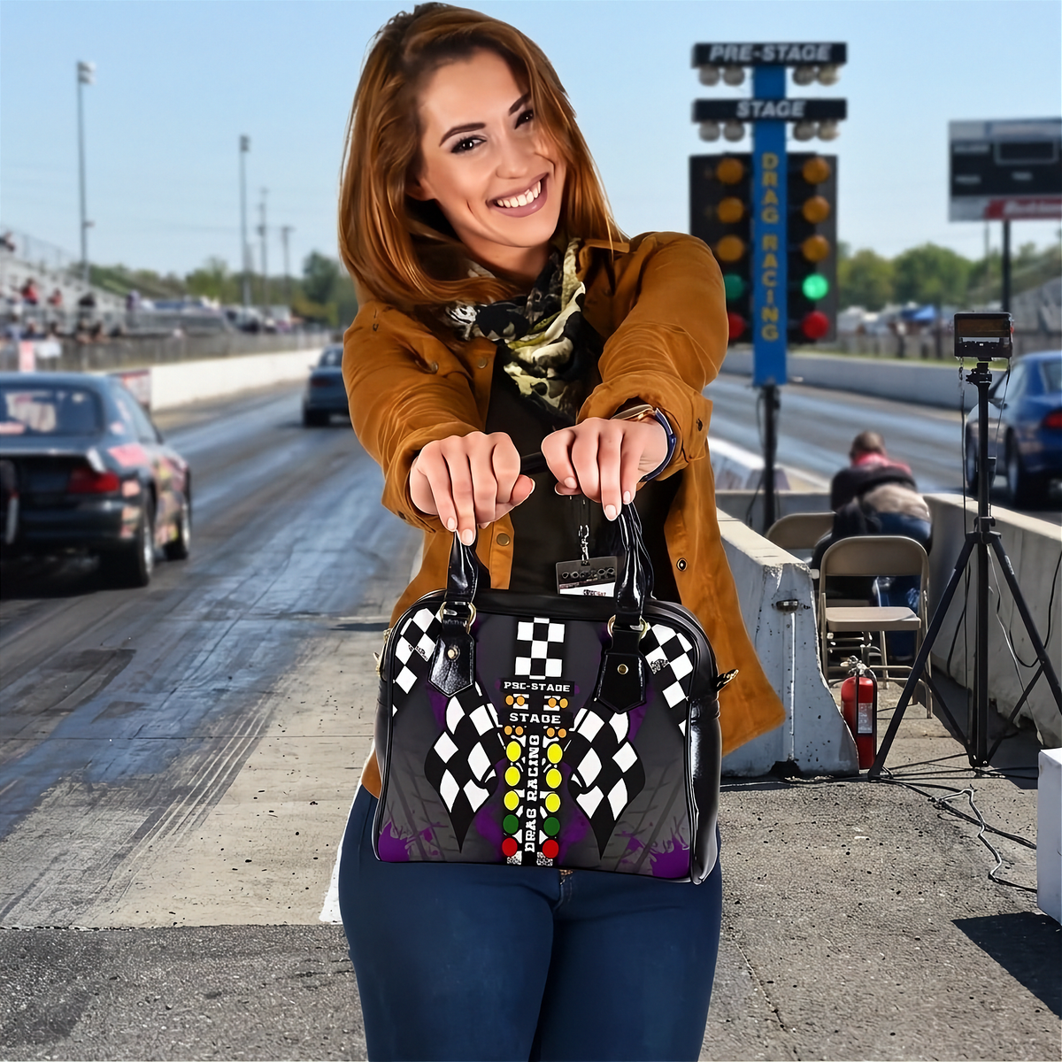 A woman at a drag racing track holds up a black handbag decorated with a checkered flag pattern and a colorful 'FIRE DEPARTMENT' graphic.