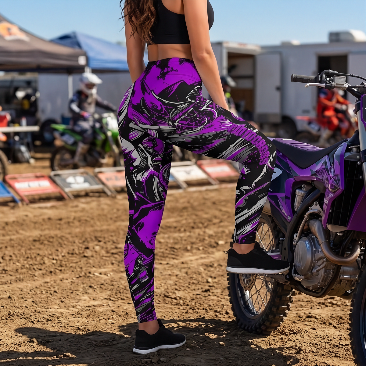 A woman wearing high-waisted purple, black, and white abstract patterned leggings is posed next to a purple and black dirt bike on a dirt track. She is wearing a black sports bra and black sneakers.