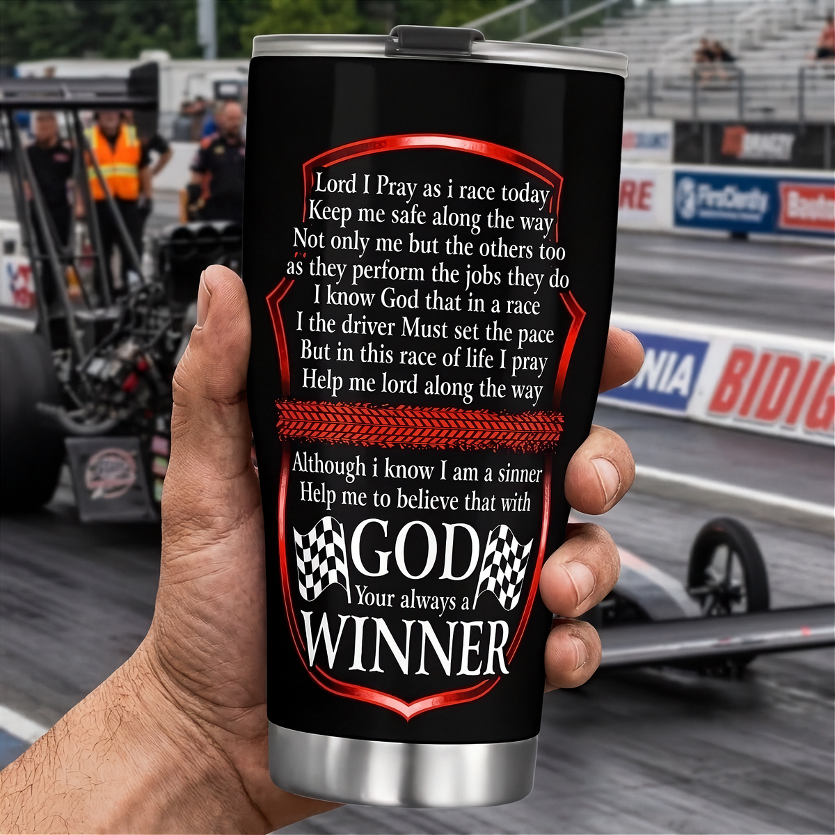 A hand holds a black insulated travel tumbler with a silver lid and base, printed with a racing-themed prayer and the phrase 'GOD Your always a WINNER' with checkered flags. The background shows a blurred racetrack scene with race cars and barriers.