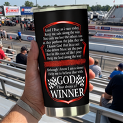 A hand holding a black insulated travel tumbler with a silver lid and base, printed with a racing-themed prayer and the phrase 'GOD Your always a WINNER' with checkered flags. The background shows a blurred racetrack scene with race cars and barriers.