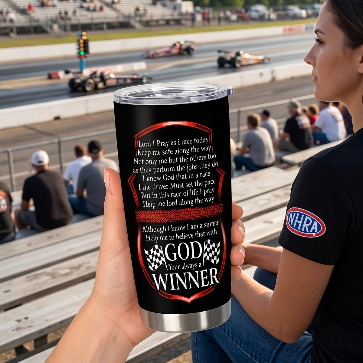 A girl holding in her hand a black insulated travel tumbler with a silver lid and base, printed with a racing-themed prayer and the phrase 'GOD Your always a WINNER' with checkered flags. The background shows a blurred racetrack scene with race cars and barriers.