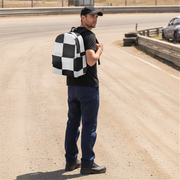 A man wearing a black t-shirt, navy blue pants, and a black cap stands on a dirt road next to a racetrack, carrying a black and white checkered backpack.